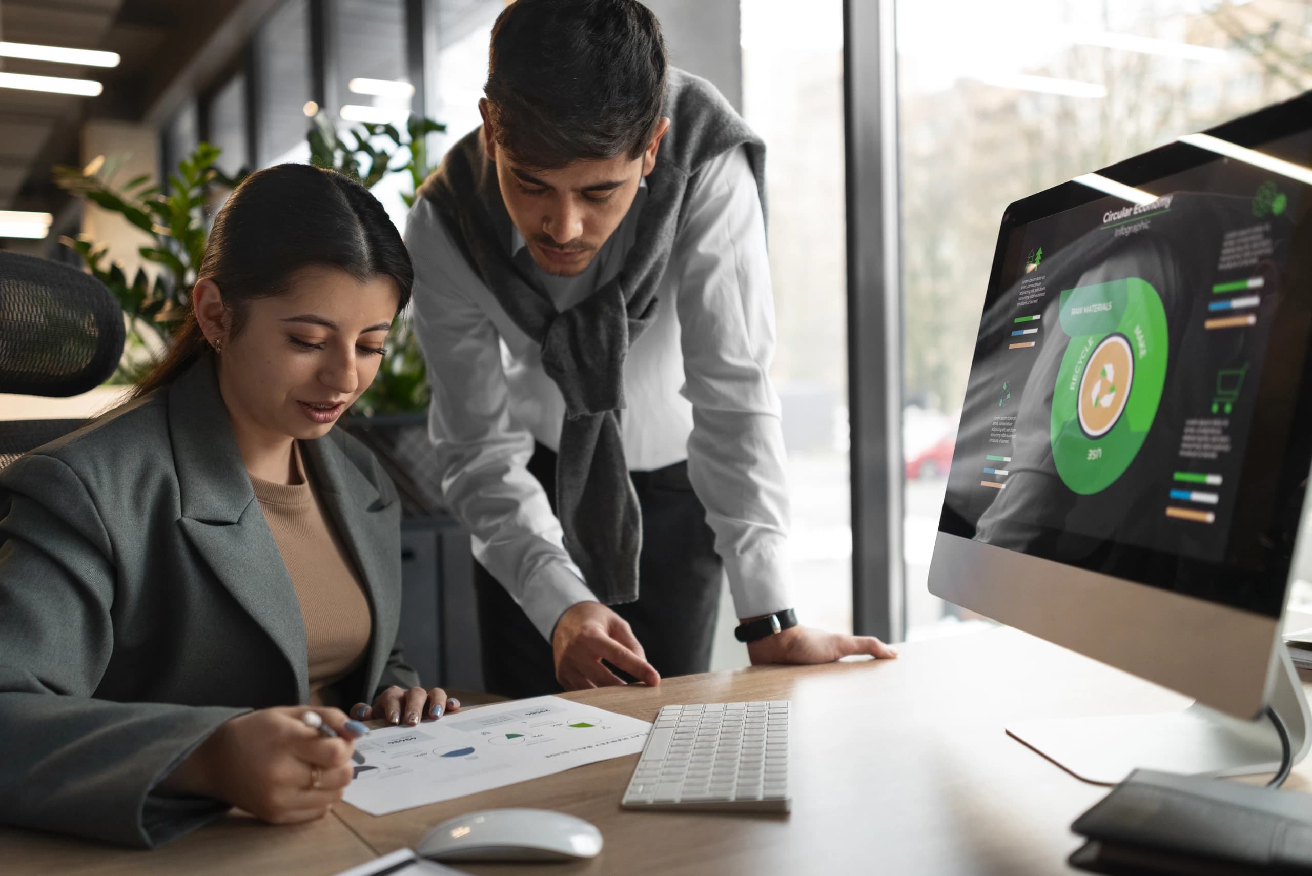 Two colleagues reviewing printed reports at a desk beside a monitor showing an analytics dashboard