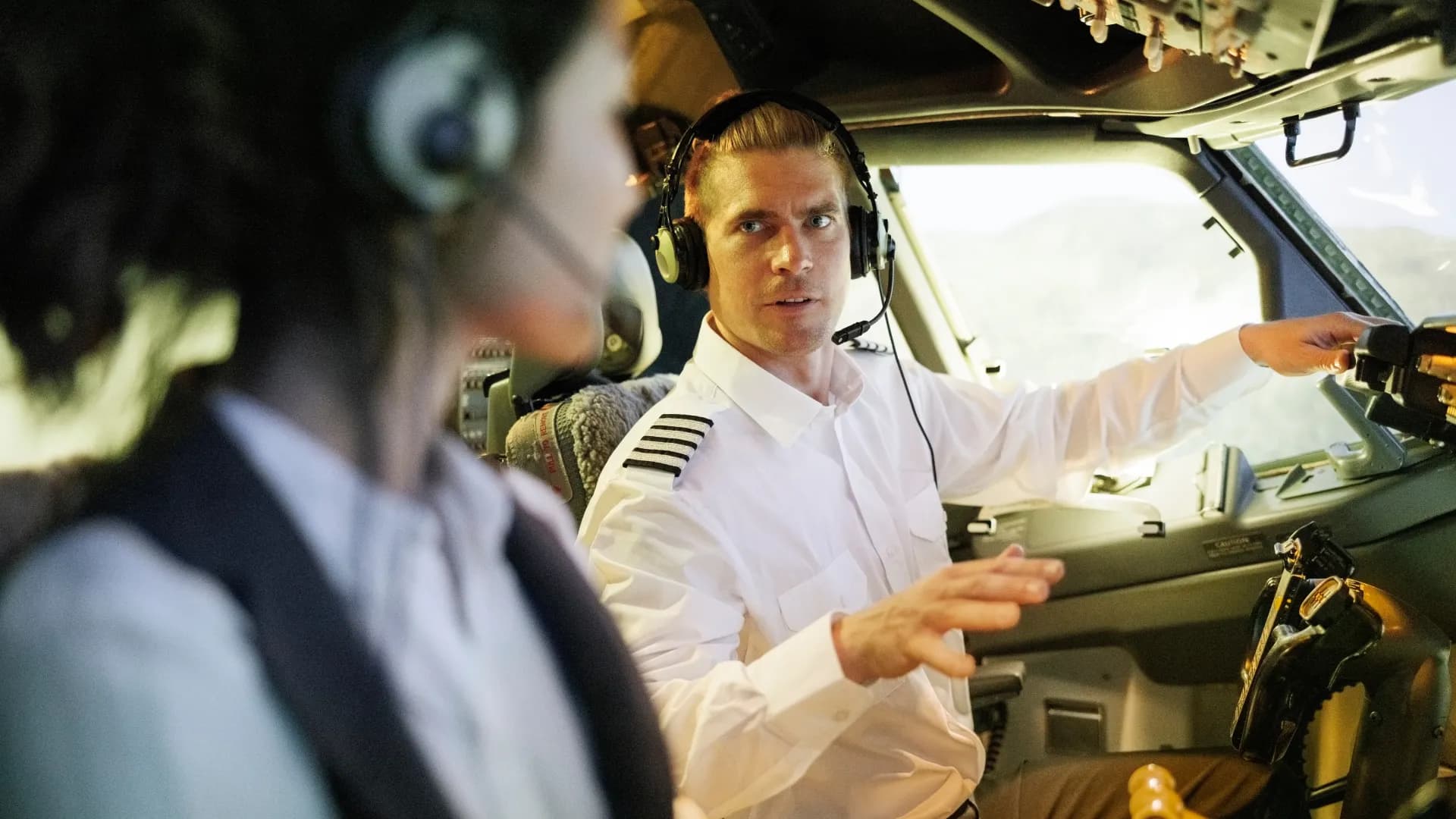 An airline captain in a headset working the flight-deck controls while talking to a crew member beside him