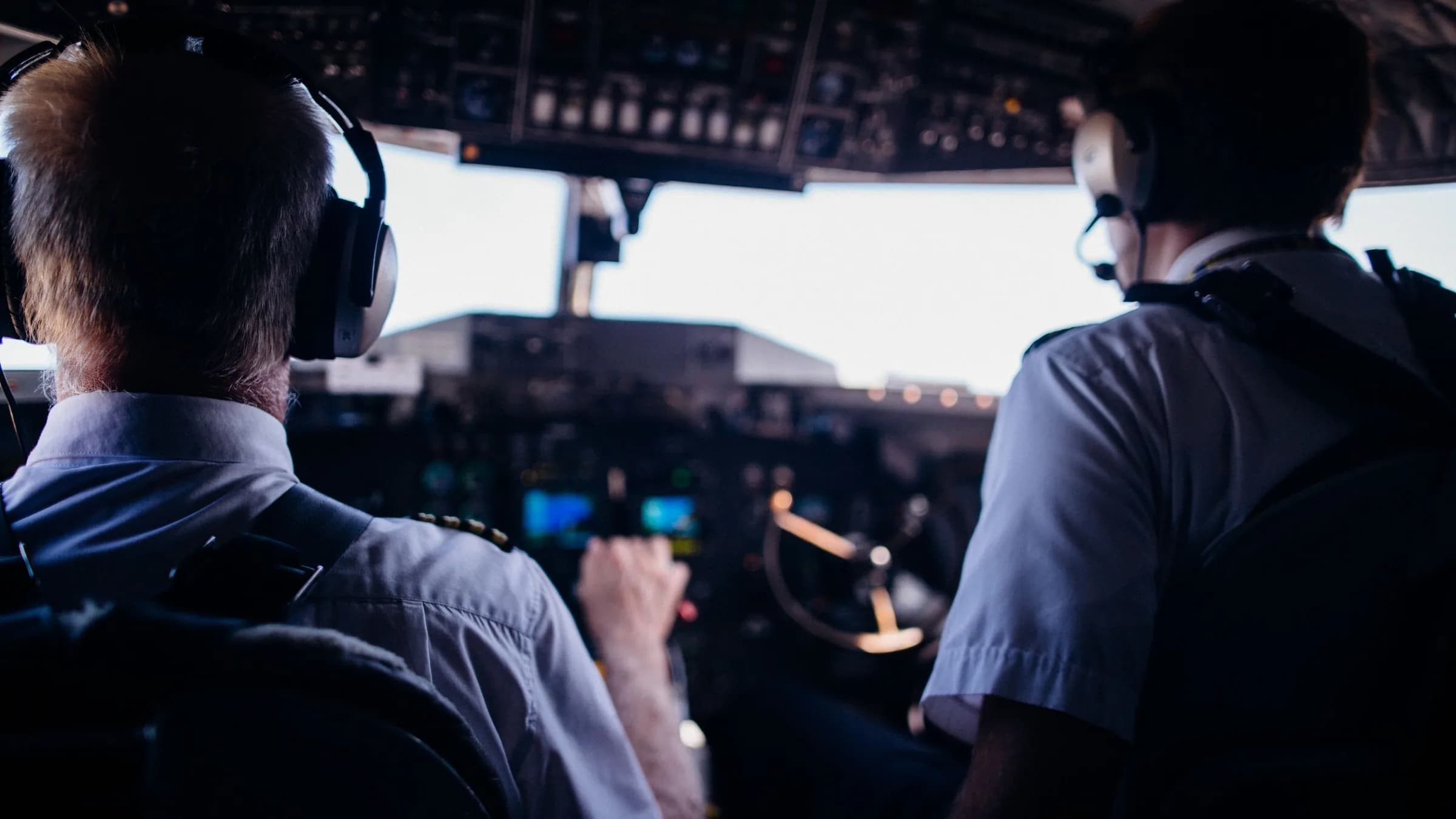 A captain and a first officer in headsets at the controls of an airliner, seen from behind on the flight deck, the instrument panels lit in front of them