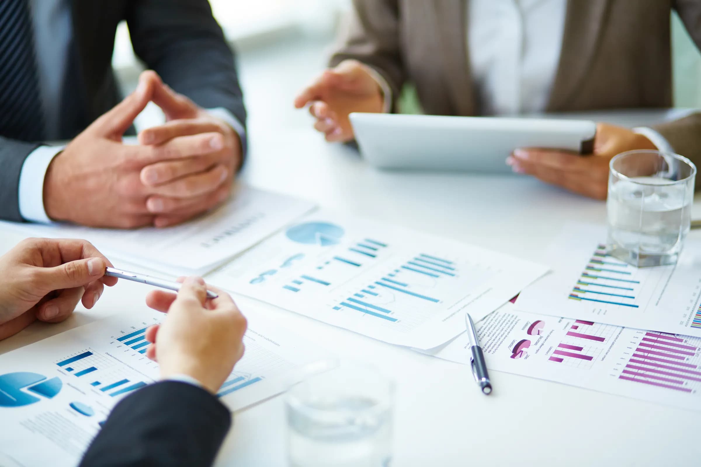 Colleagues around a meeting table reviewing printed bar and pie charts, one holding a pen over the reports while another holds a tablet