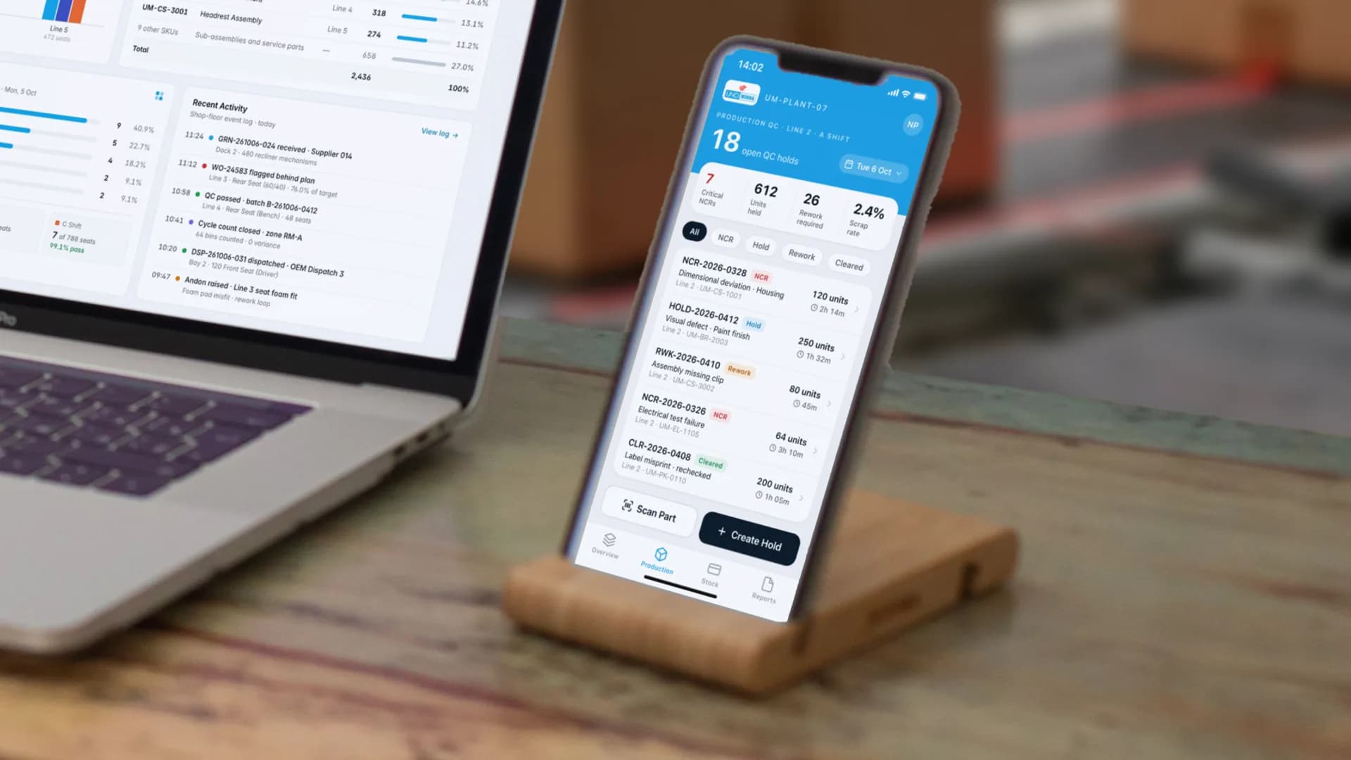 The QC Holds screen on an iPhone in a stand on a desk by the line, Line 2 on A Shift: 7 critical NCRs, 612 units held, 26 units requiring rework and a 2.4% scrap rate across 18 open holds, over a list filtered by NCR, hold, rework and cleared with the units and the time held on each, and Scan Part and Create Hold actions