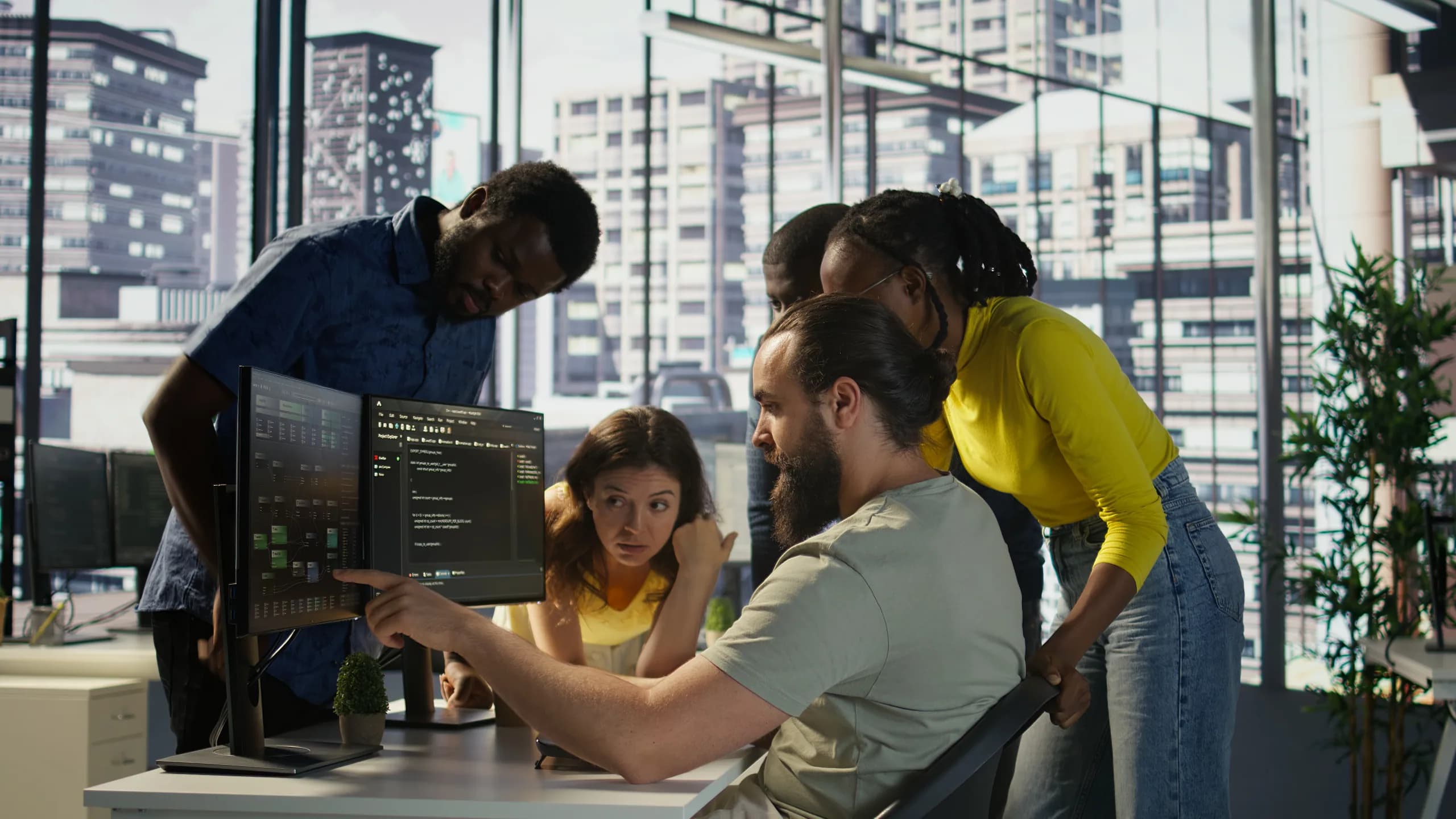 Five colleagues gathered around a desk as one points at code on a pair of monitors