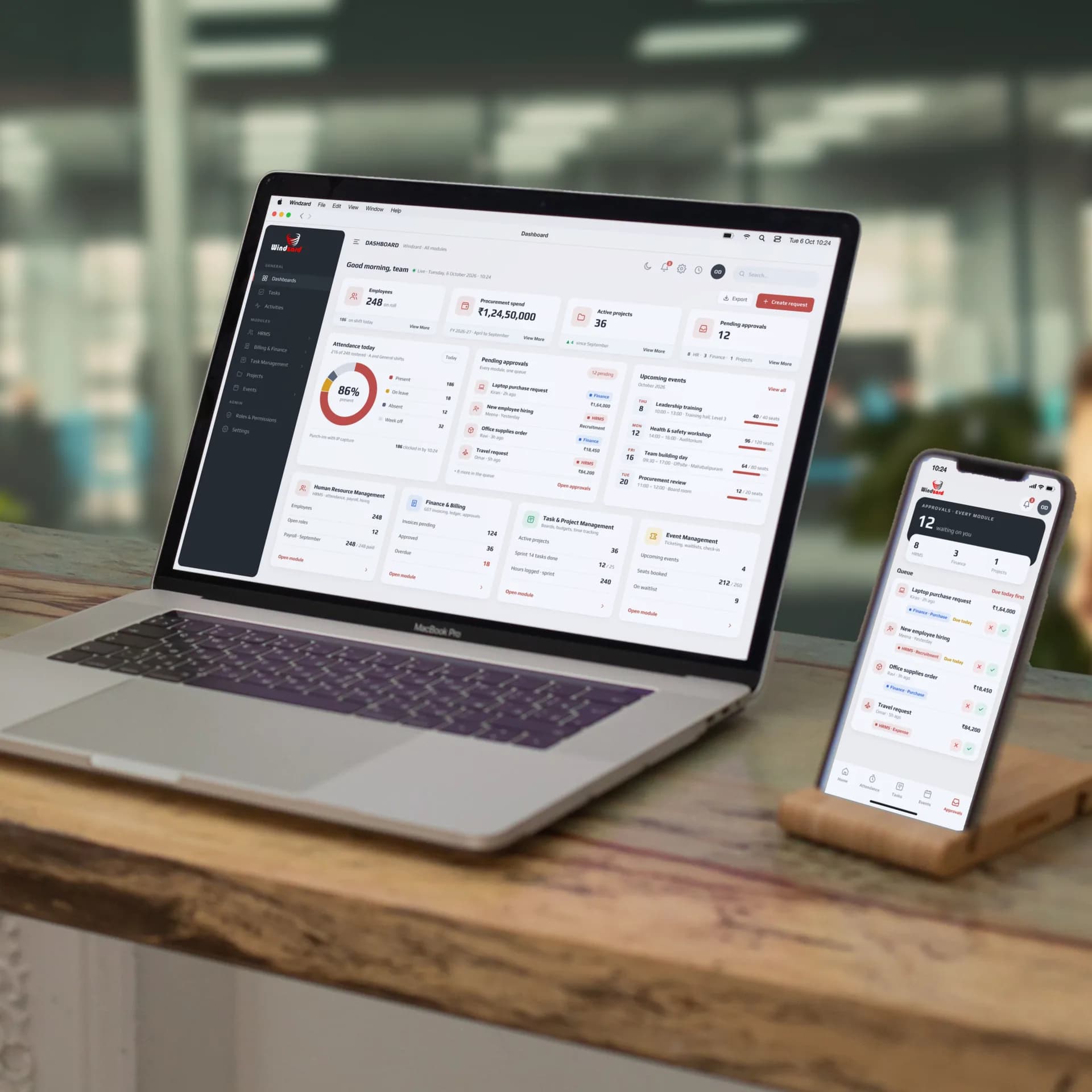 The Windzard enterprise platform we built, on a MacBook Pro and an iPhone in a stand on a desk in an open-plan office: the dashboard with 248 employees, procurement spend, active projects and pending approvals, and the approvals queue on the phone