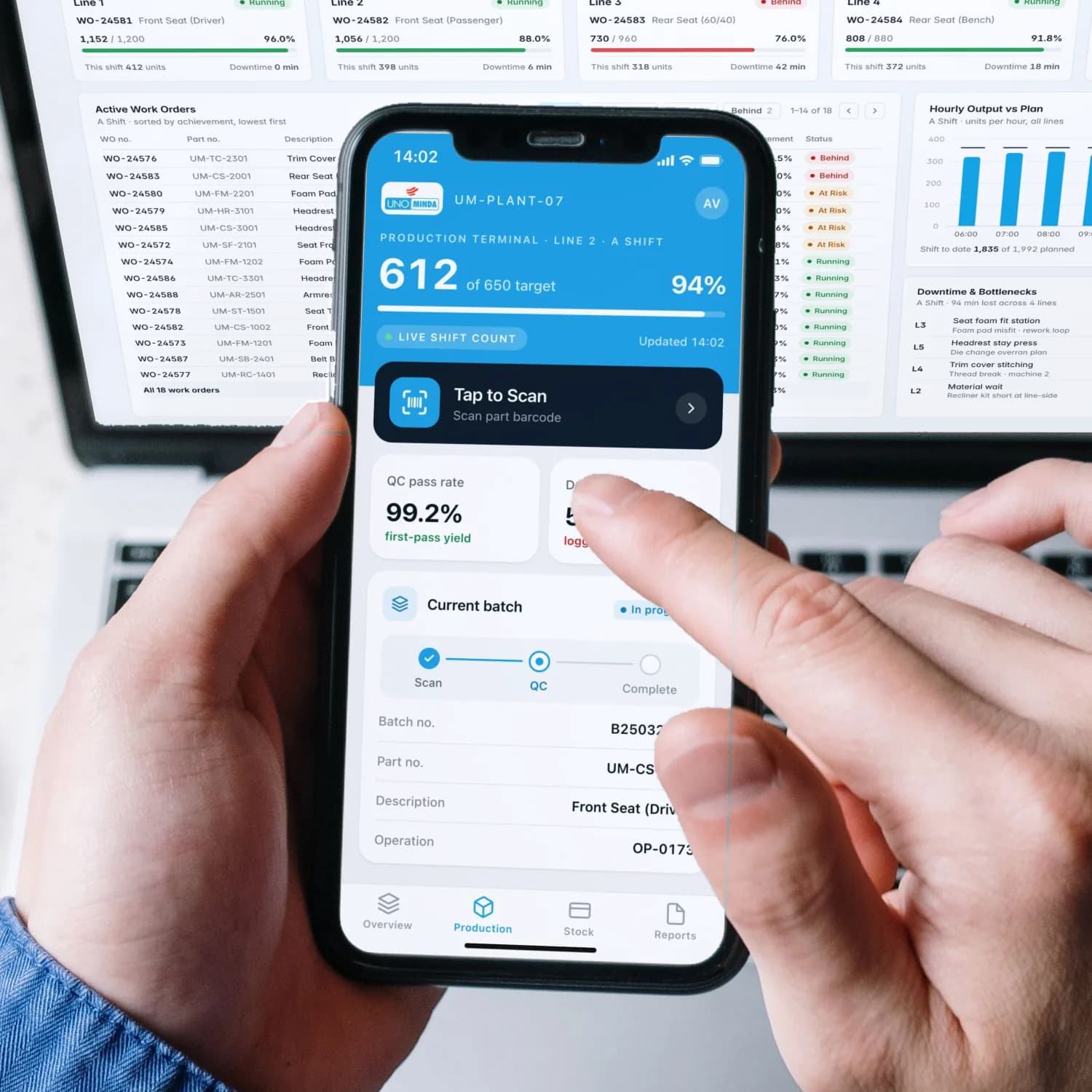 Two hands holding an iPhone running the UNO Minda production terminal app we built — a live shift count of 612 of a 650 target and a Tap to Scan action — over a MacBook Pro showing the production console