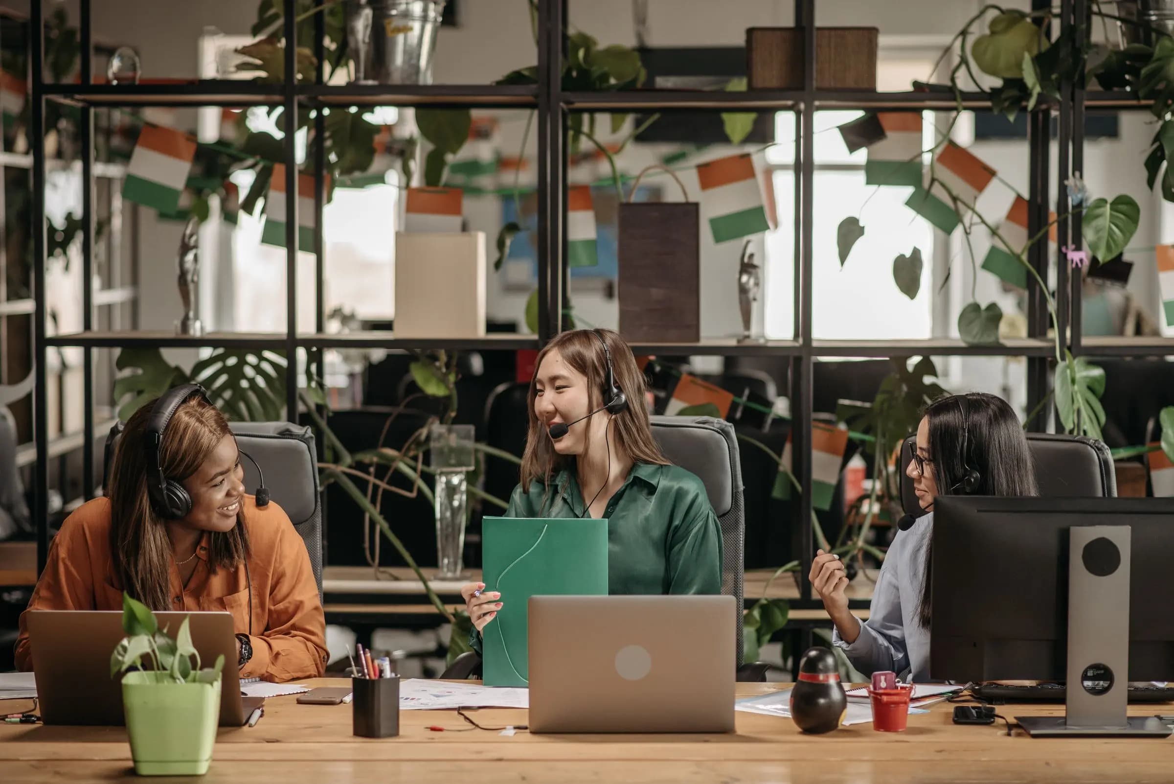 Three women wearing headsets talk and laugh across a shared desk with laptops in a plant-filled office.