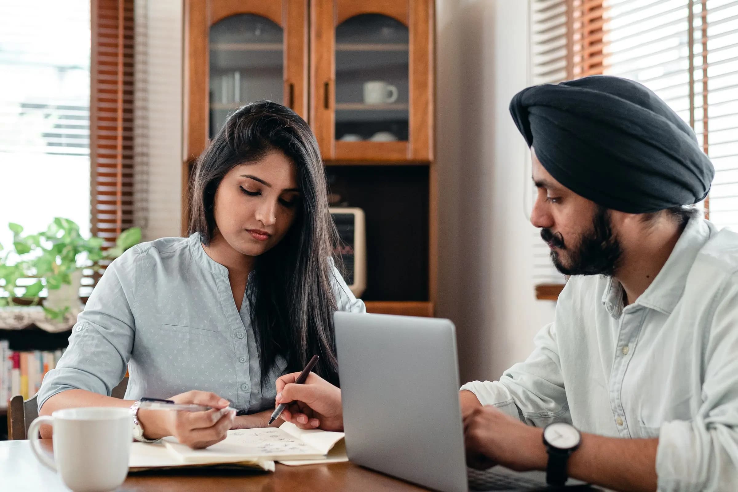 A man and a woman review figures together at a table, one writing in a notebook and the other working on a laptop.