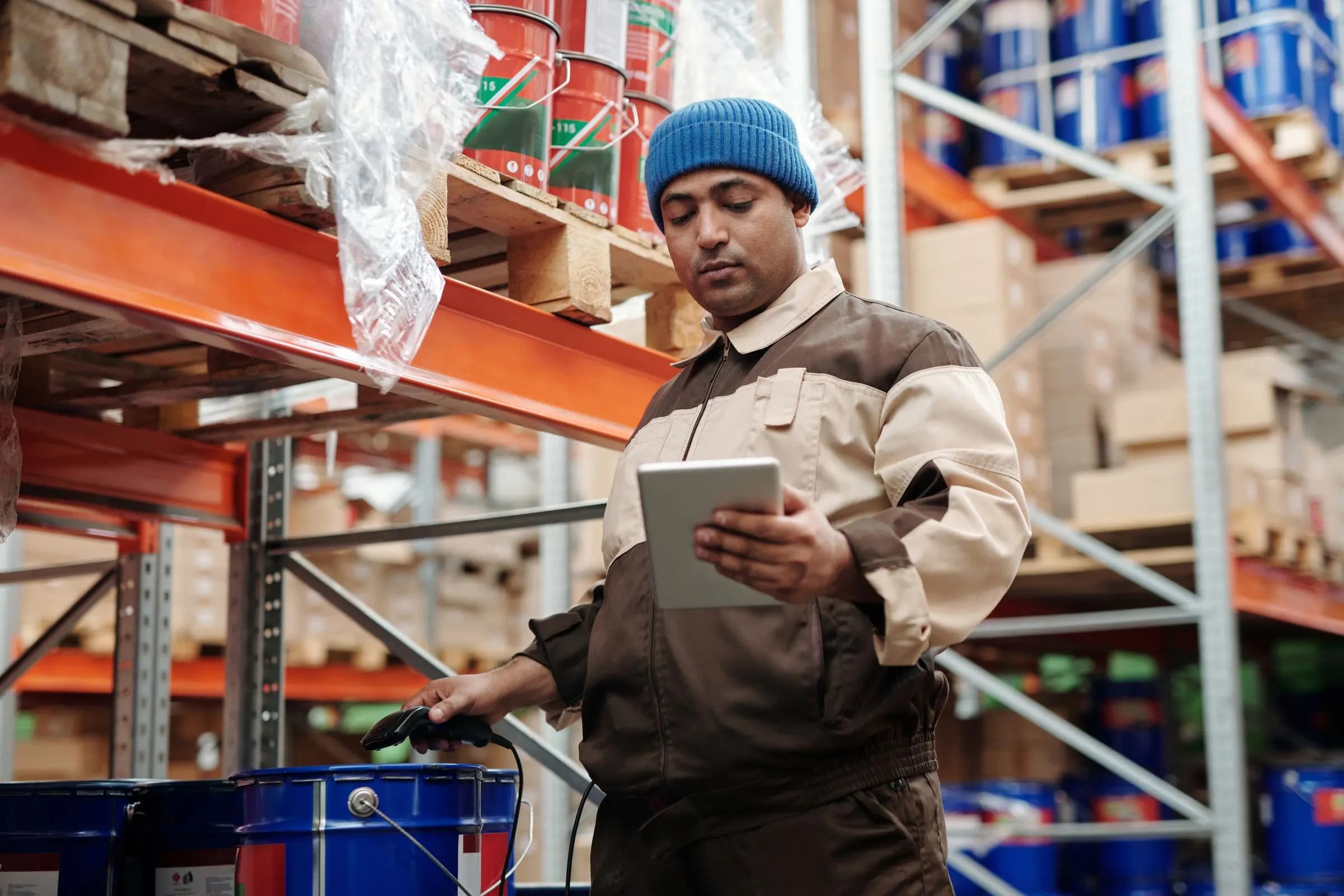 A warehouse worker in a knit cap holds a tablet and a barcode scanner beside shelves of stock.