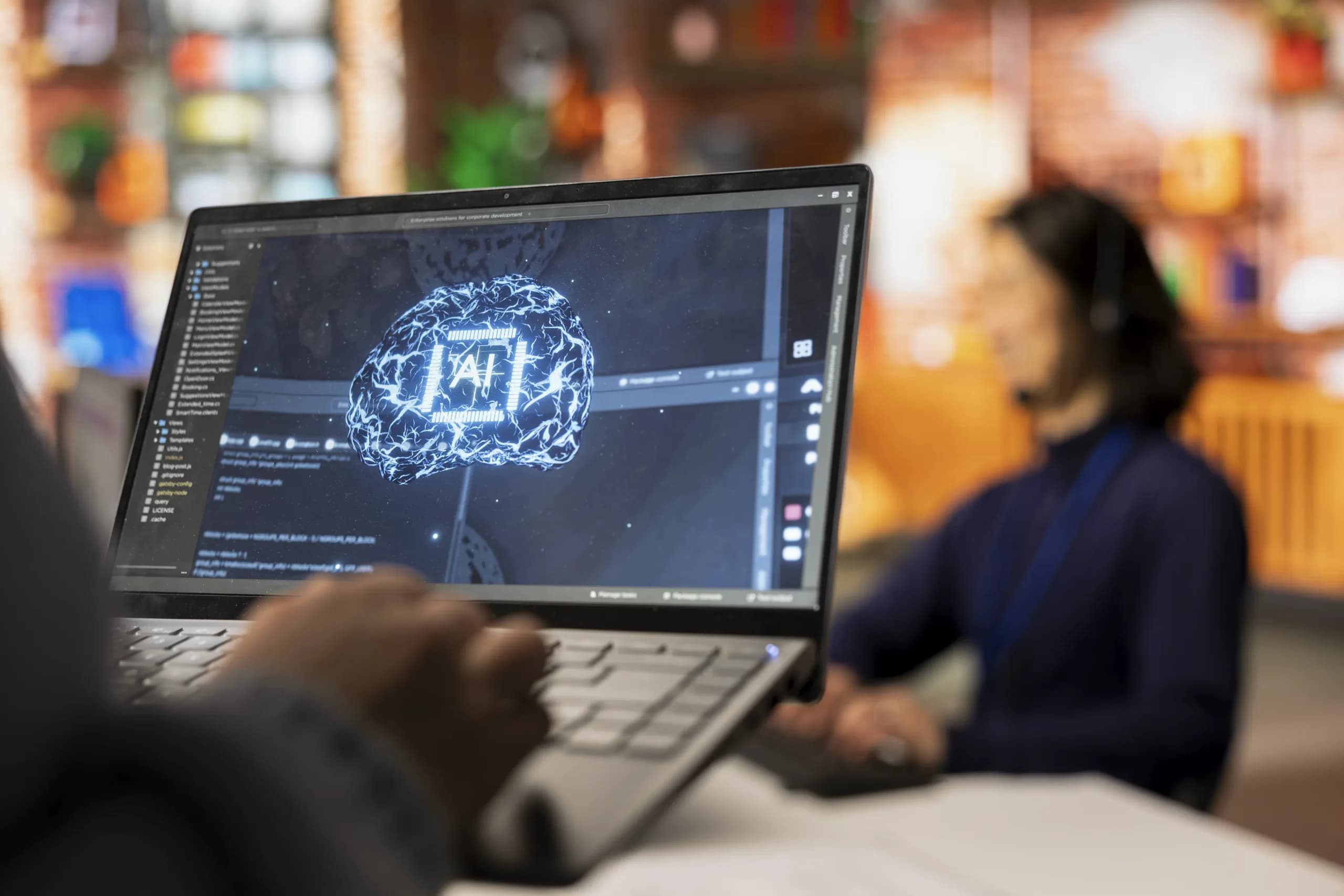 A developer working on an AI model on a laptop, a colleague at a second screen behind
