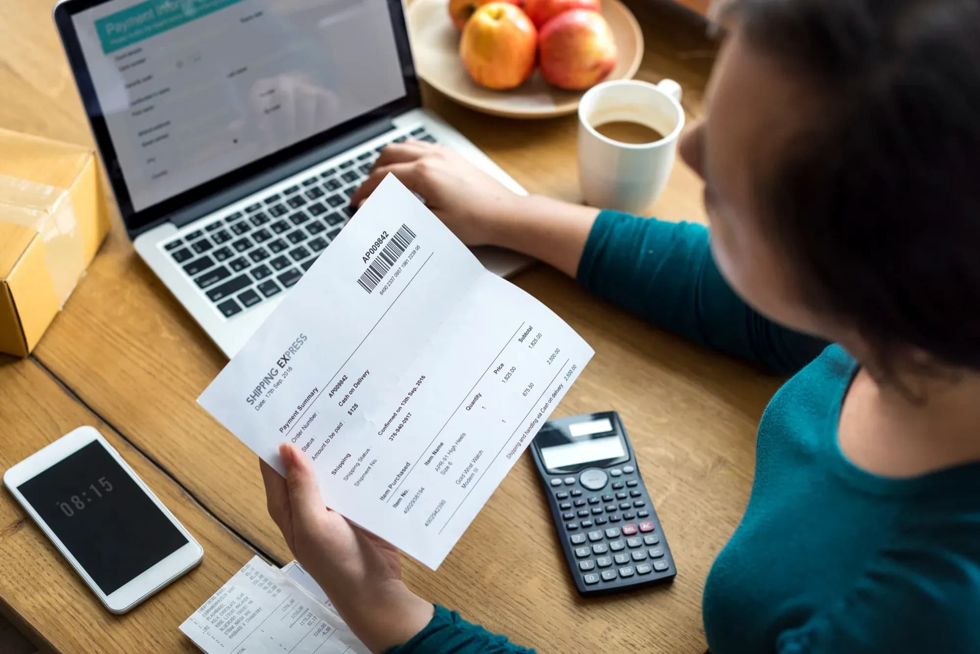 Someone at a laptop checking a printed payment summary against the payment form on screen, a calculator and receipts beside them