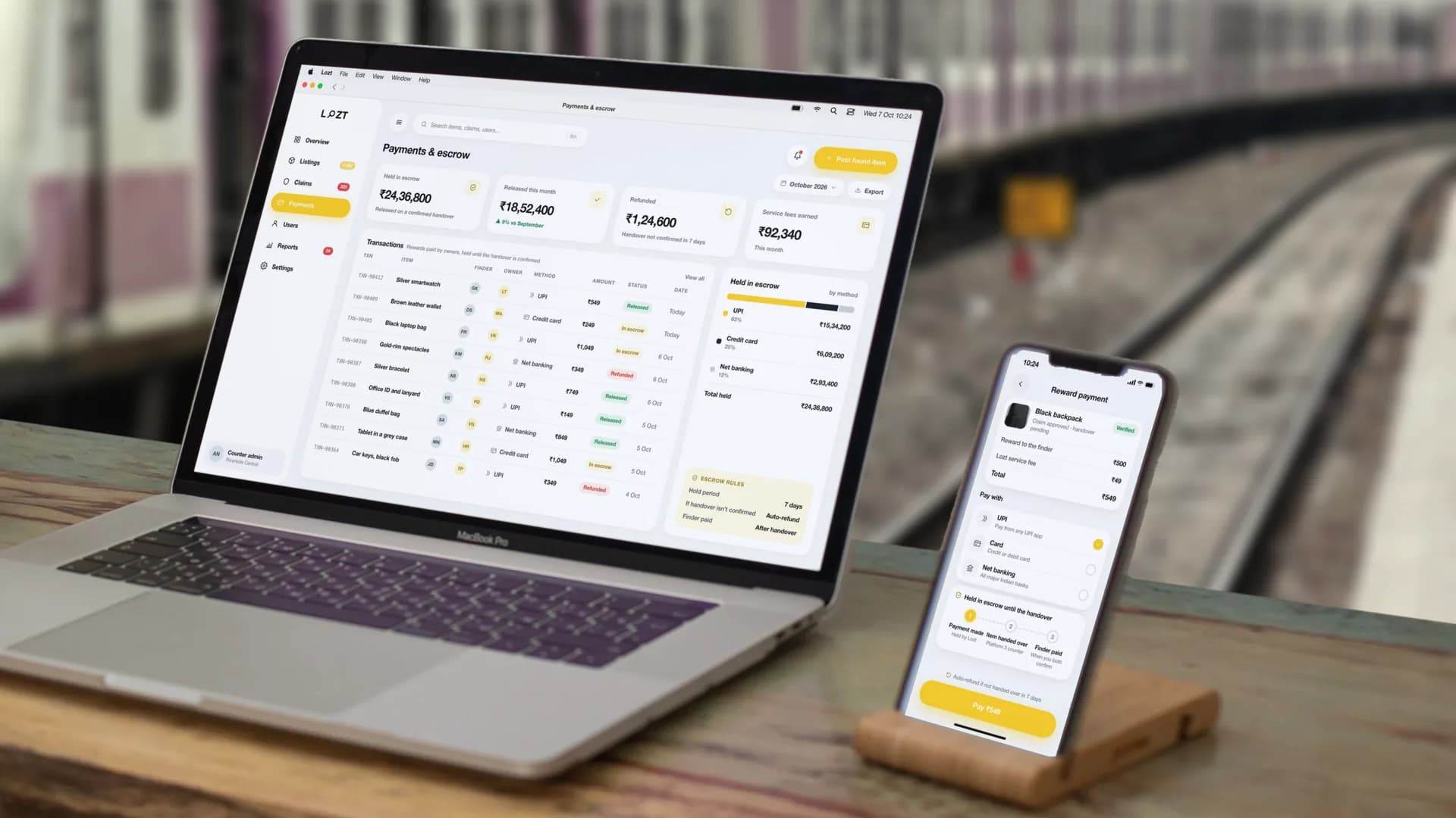 The payments console on a MacBook Pro, the app’s reward payment on an iPhone beside it, on a desk on a railway platform: ₹24,36,800 held in escrow, ₹18,52,400 released this month, ₹1,24,600 refunded and ₹92,340 in service fees earned, above a transaction ledger carrying the item, the finder, the owner, the method — UPI, credit card or net banking — and whether each sits In escrow, Released or Refunded, beside what is held split by method