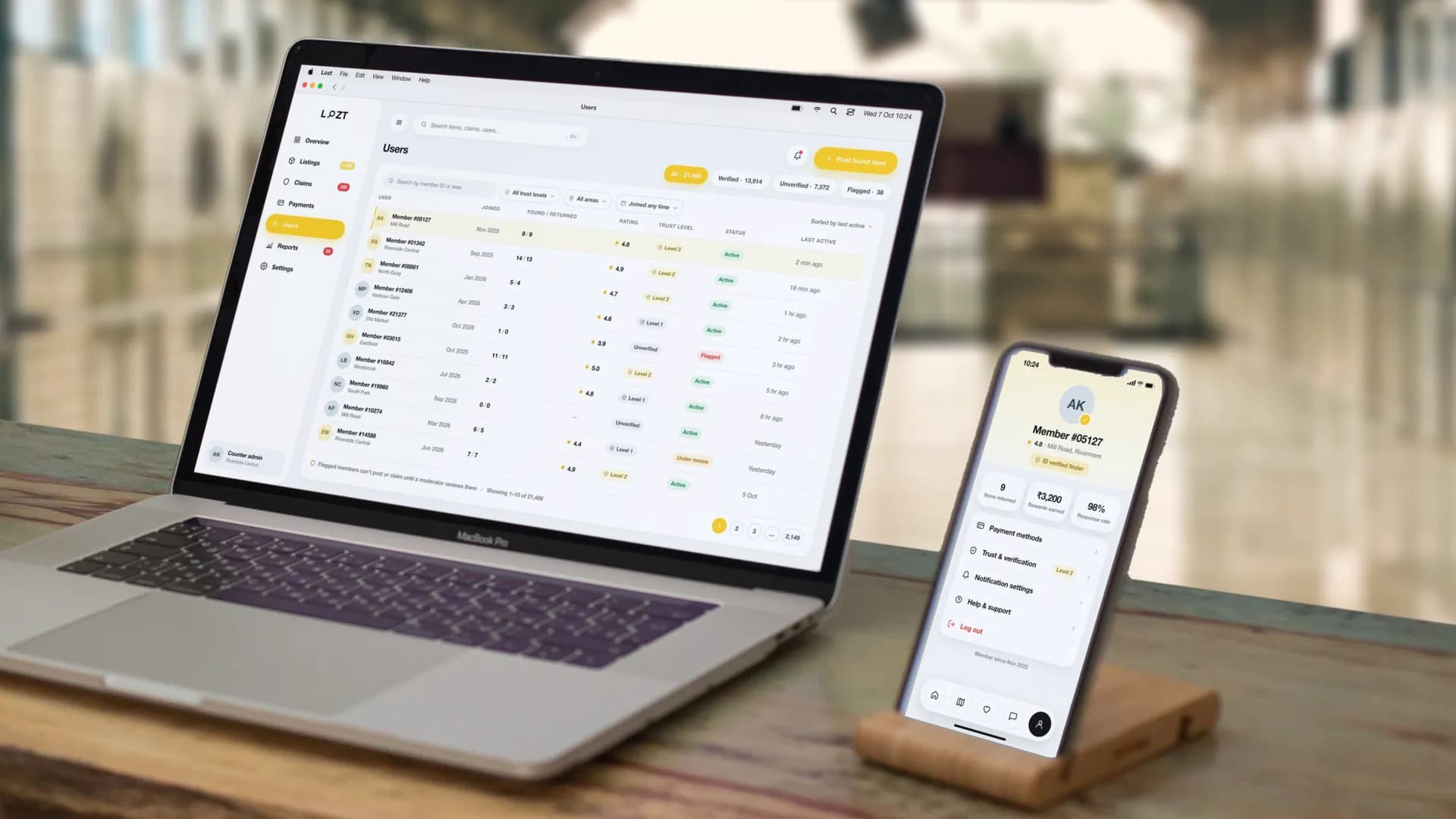 The Users console: 21,486 members tabbed 13,914 verified, 7,572 unverified and 38 flagged, every member shown only by initials and a member number, with their area, the month they joined, items found and returned, rating, trust level — Level 1, Level 2 or unverified — and an Active, Flagged or Under review status — on a MacBook Pro beside the app’s Profile screen on an iPhone, on a desk on a station platform