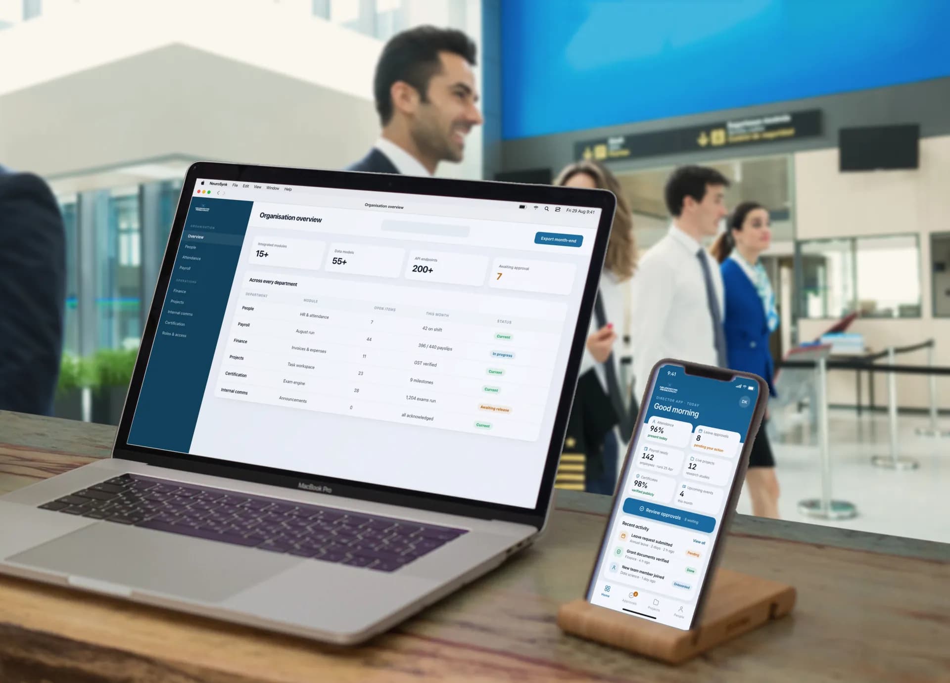 The NeuroSynk Research organisation overview on a MacBook Pro — 15+ integrated modules, 55+ data models, 200+ API endpoints and 7 items awaiting approval, with every department's open items and status — beside the Director App home on an iPhone, on a desk with airline pilots and a cabin crew member walking through an airport terminal behind