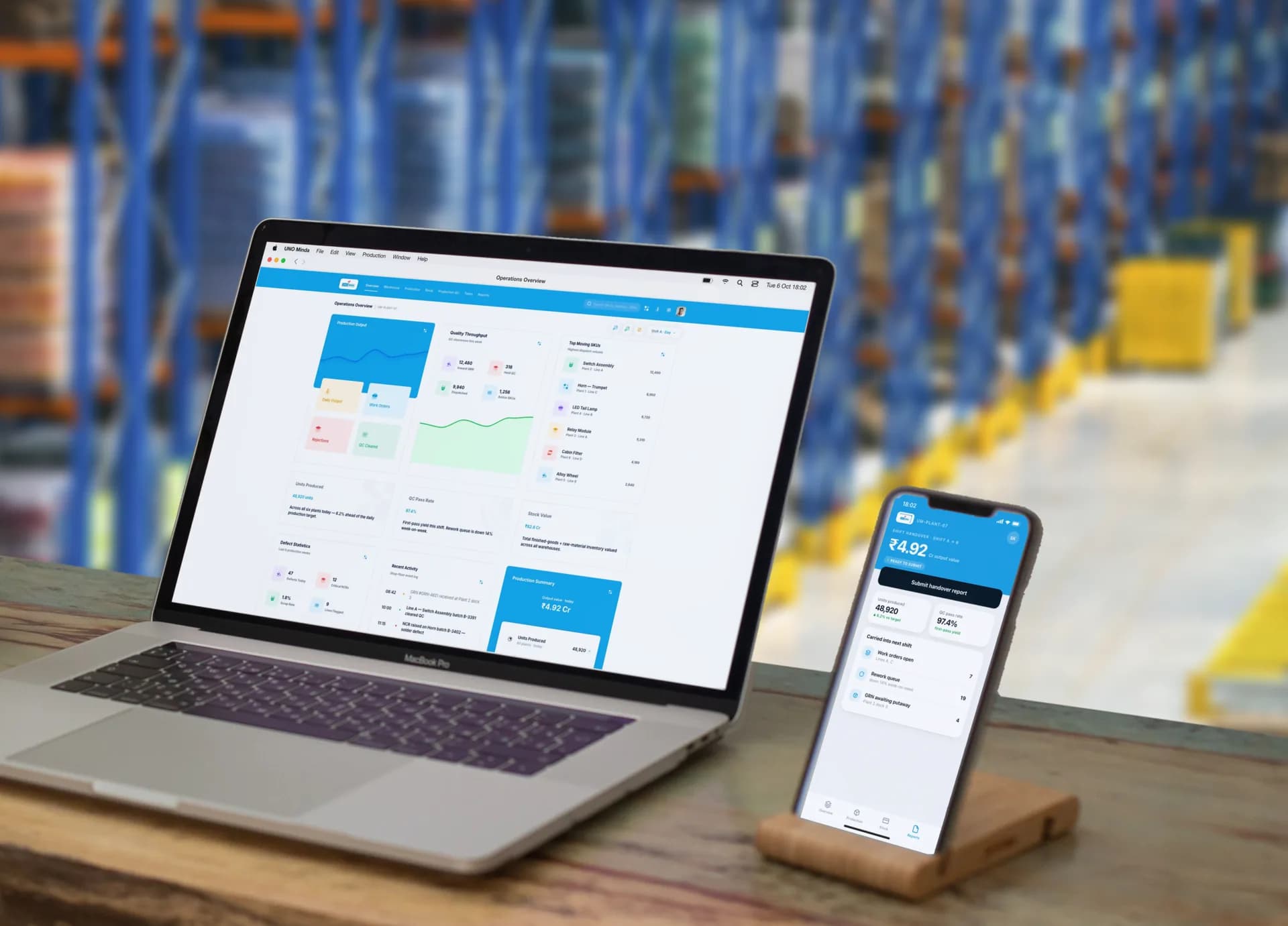 The UNO Minda operations overview on a MacBook Pro beside the shift handover screen on an iPhone in a stand, on a desk at the end of a warehouse aisle, both showing the same shift: 48,920 units produced, a 97.4% QC pass rate and ₹4.92 Cr of output