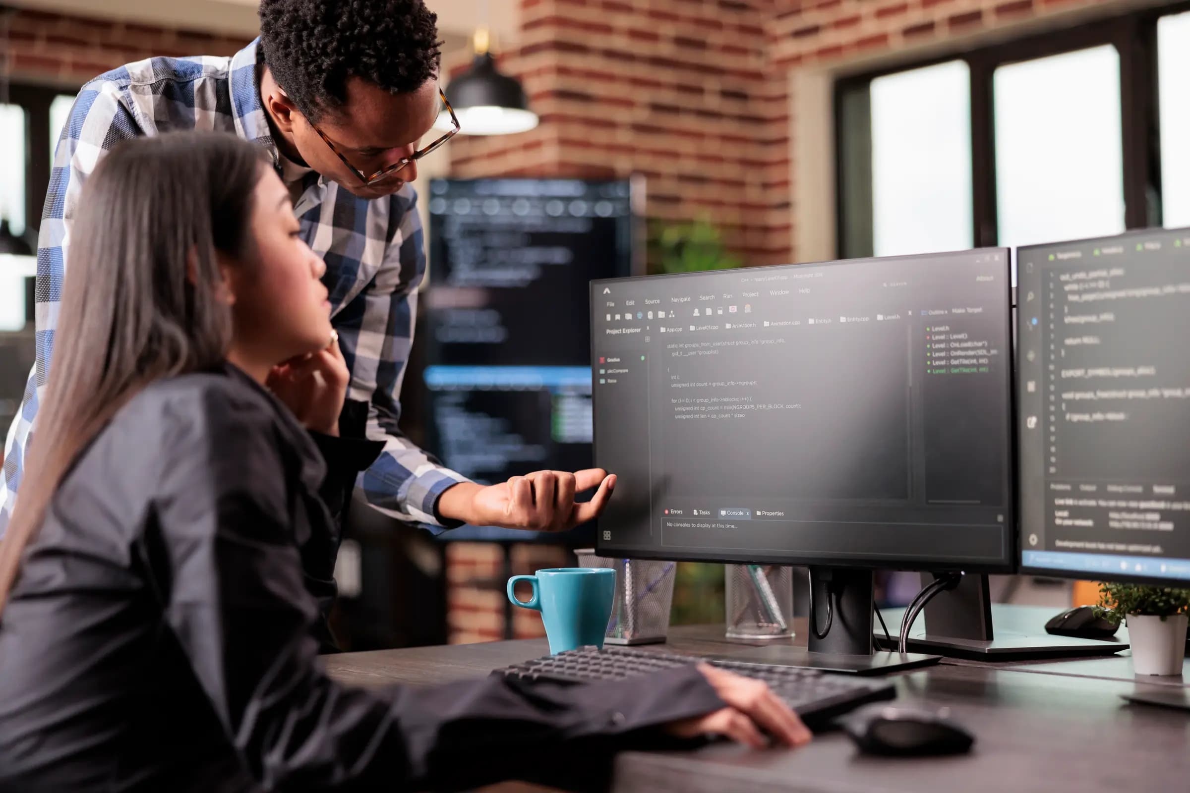 Two programmers at a desk in a brick-walled office, one standing and pointing at code on a monitor while the other, seated, follows along