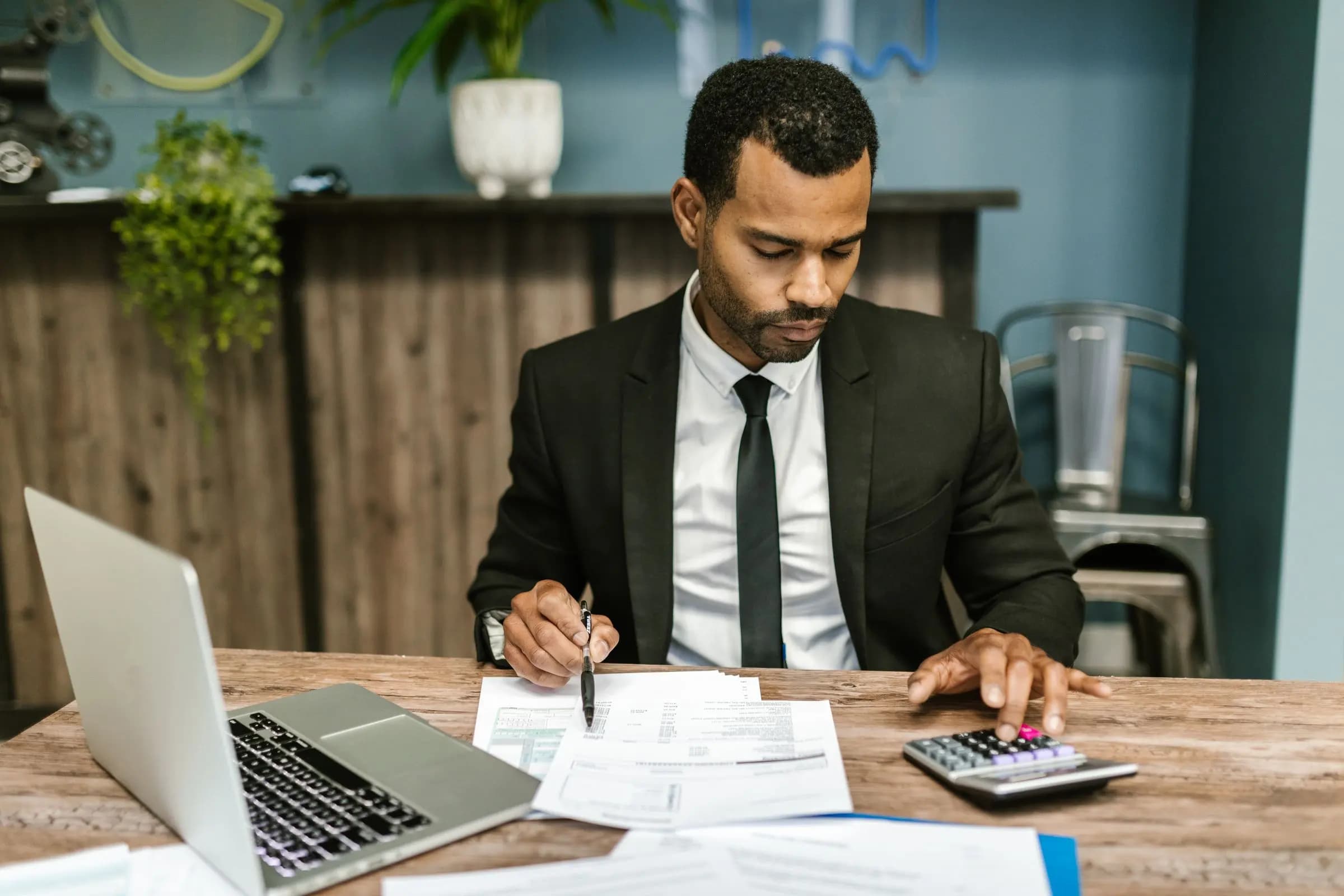 A man in a dark suit works through printed invoices with a calculator beside an open laptop.