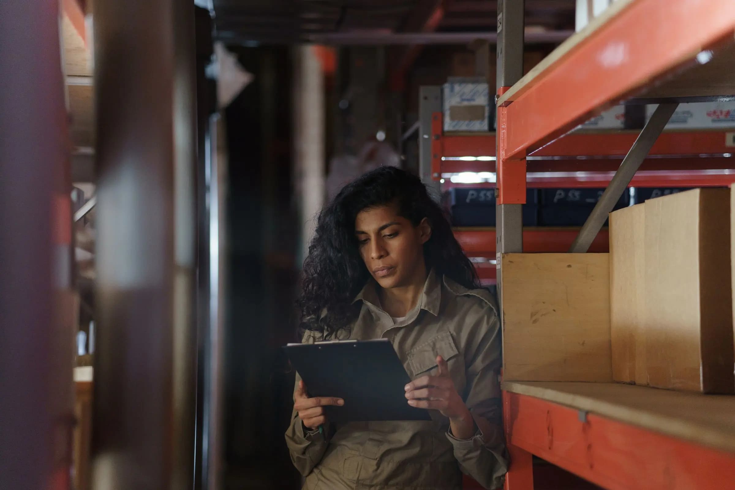 A woman checks a clipboard between orange storage shelves stacked with boxes.