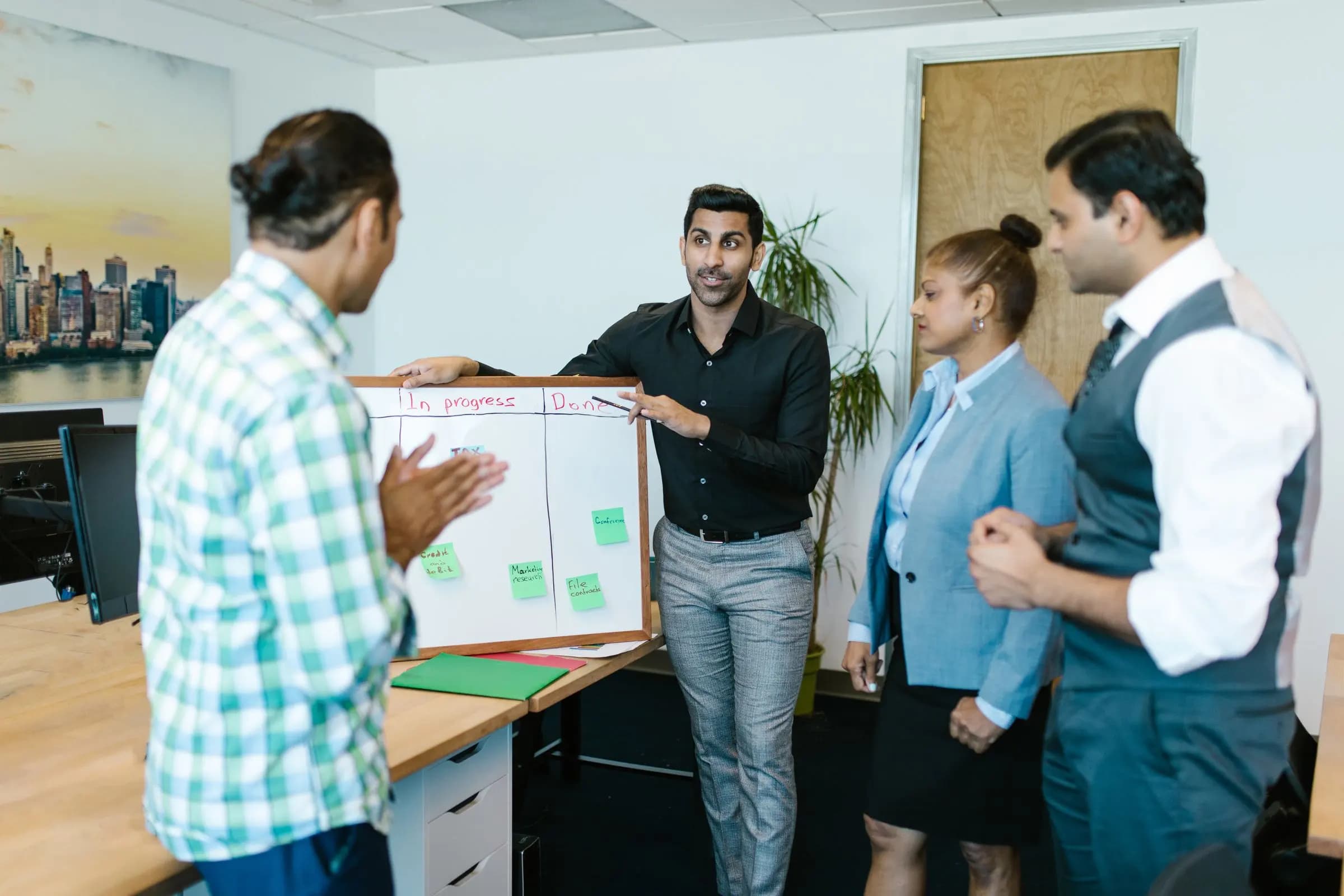 A team stands around a whiteboard with in-progress and done columns while one colleague walks through the plan.