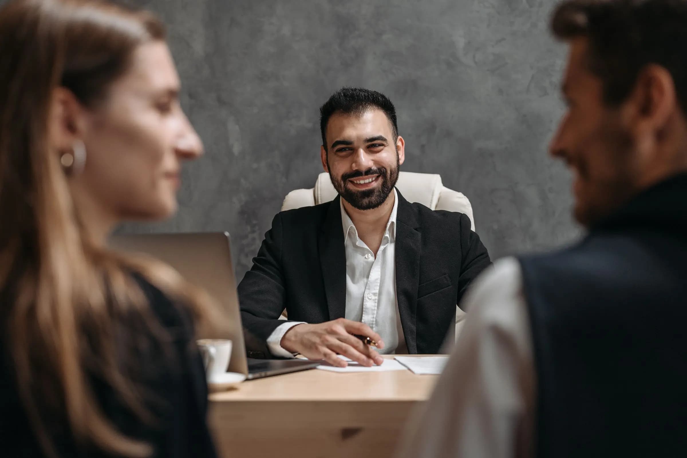 A smiling man at a desk talks with two visitors seated across from him.