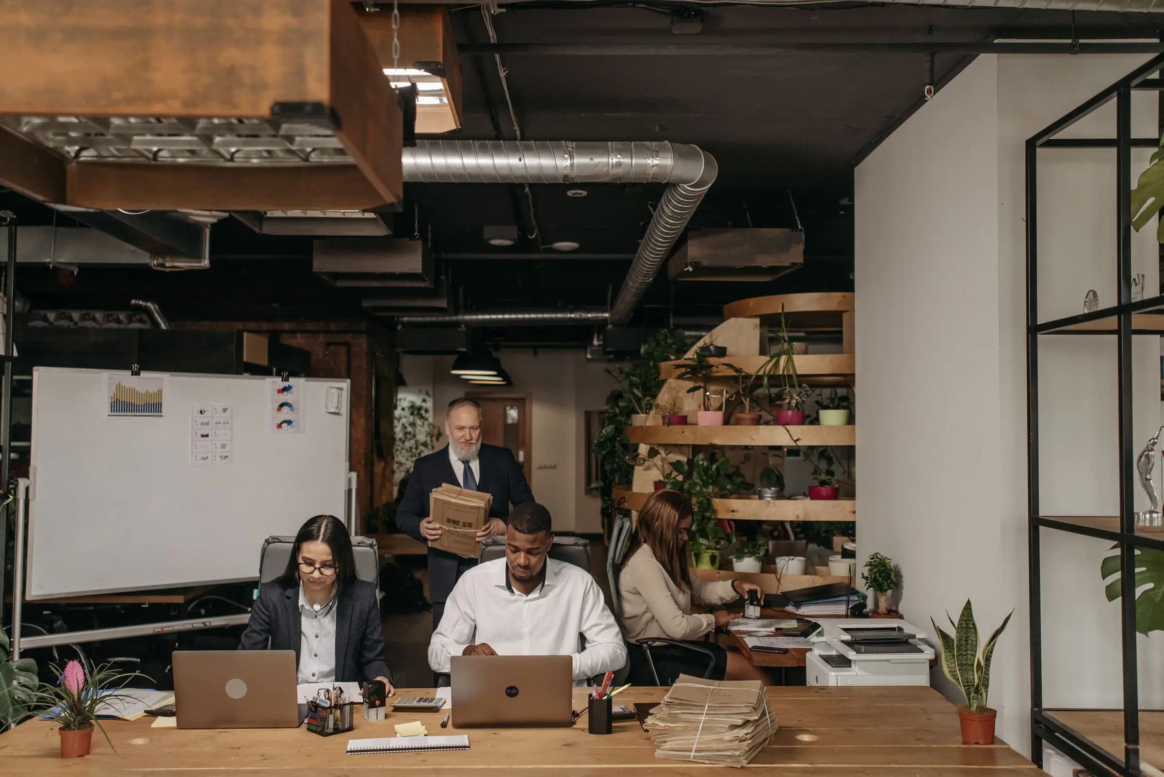 Employees work at laptops and paperwork in an open office while a colleague carries files behind them.