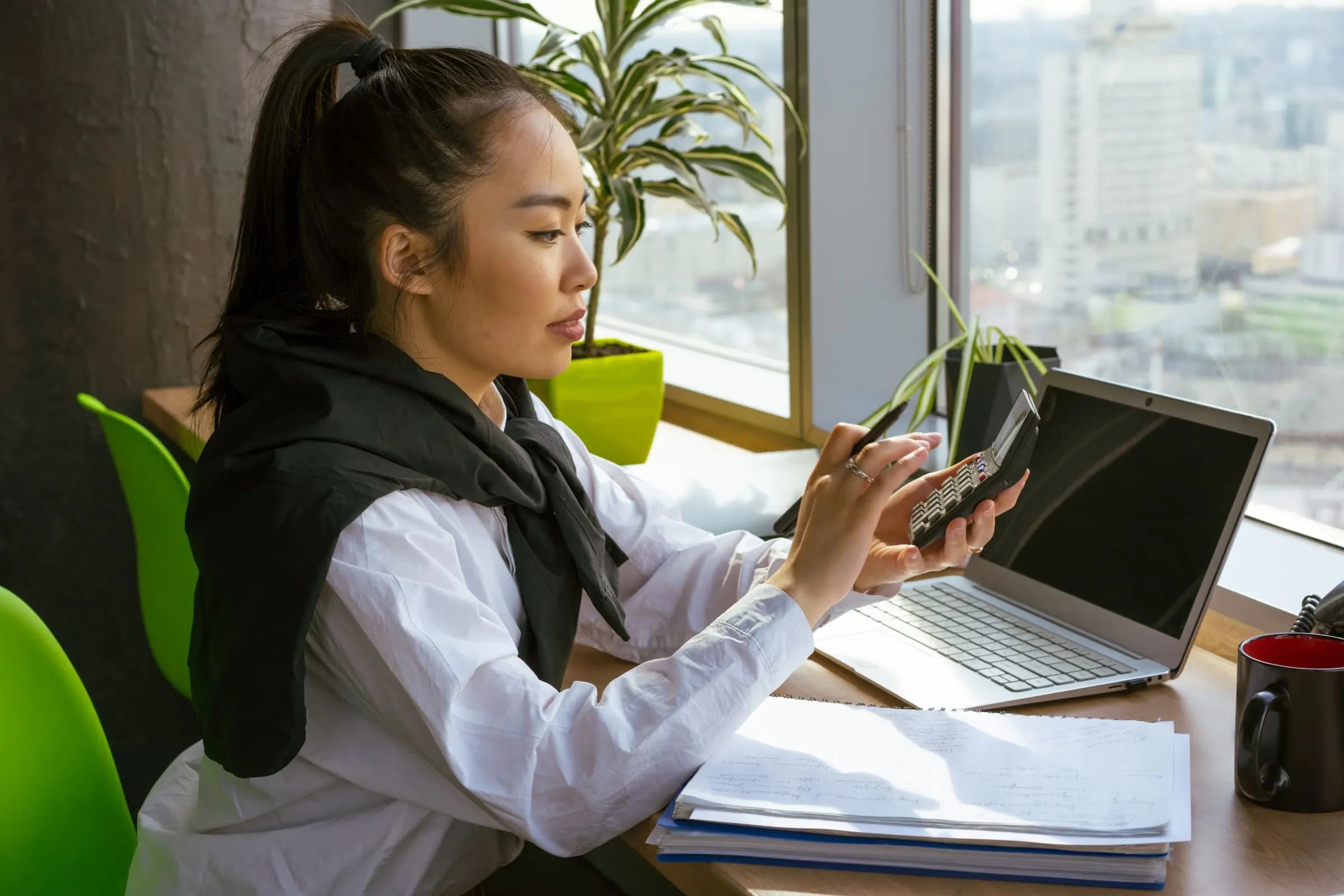 A woman by a window works out figures on a calculator next to a laptop and a stack of papers.