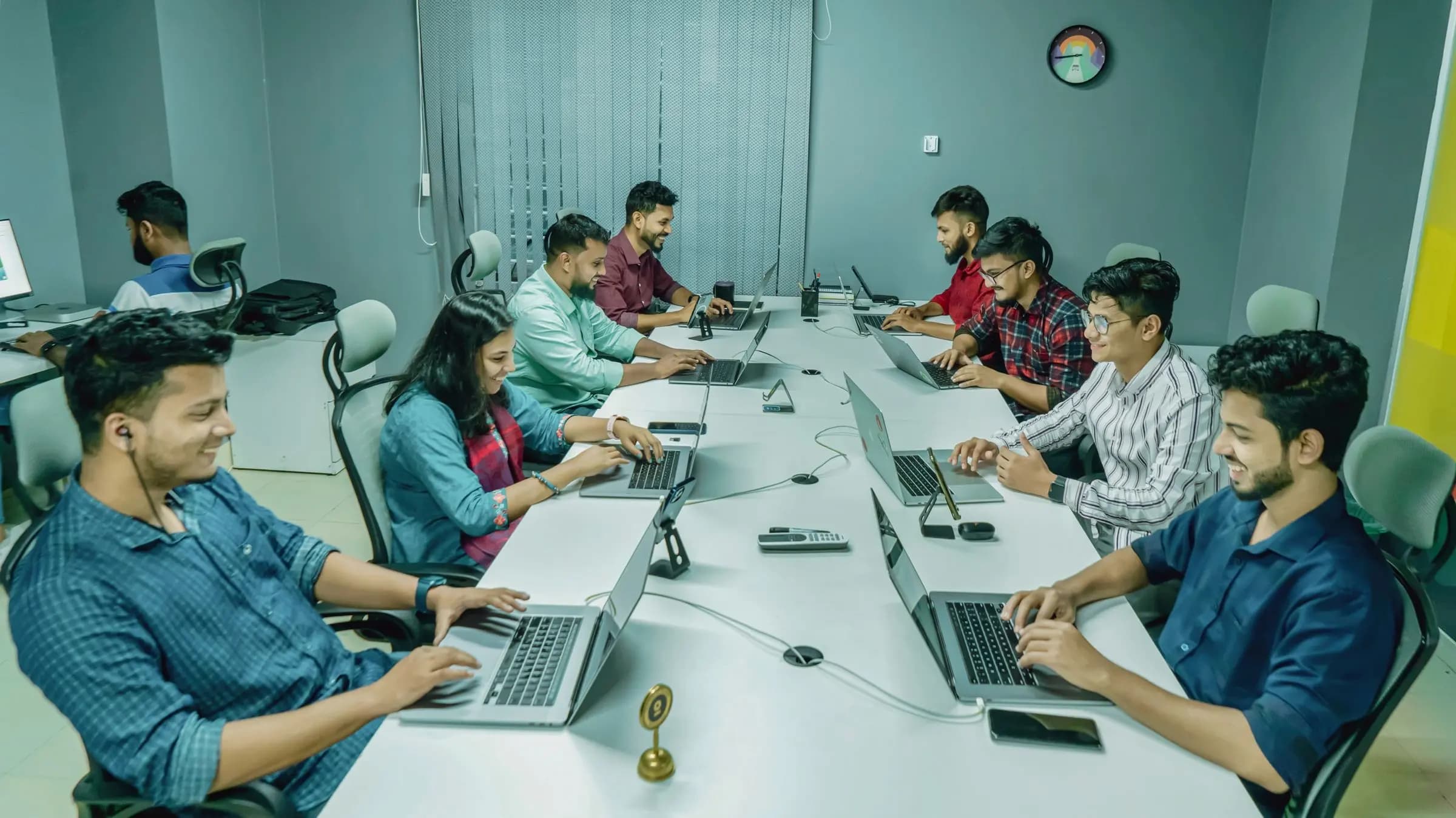A team of young colleagues works on laptops around a long shared table in an office.