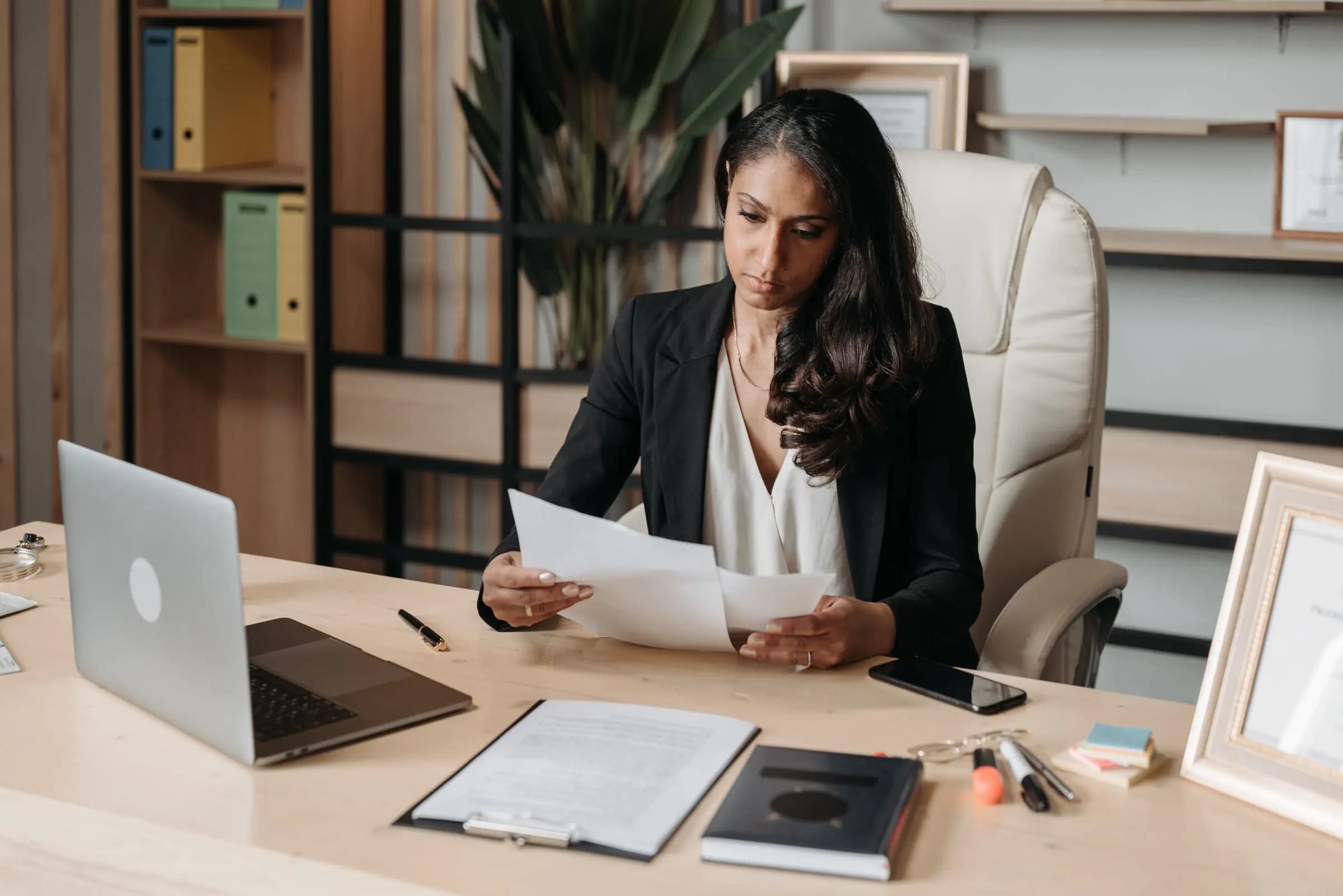 A woman at an office desk reads through printed documents next to an open laptop and a clipboard.