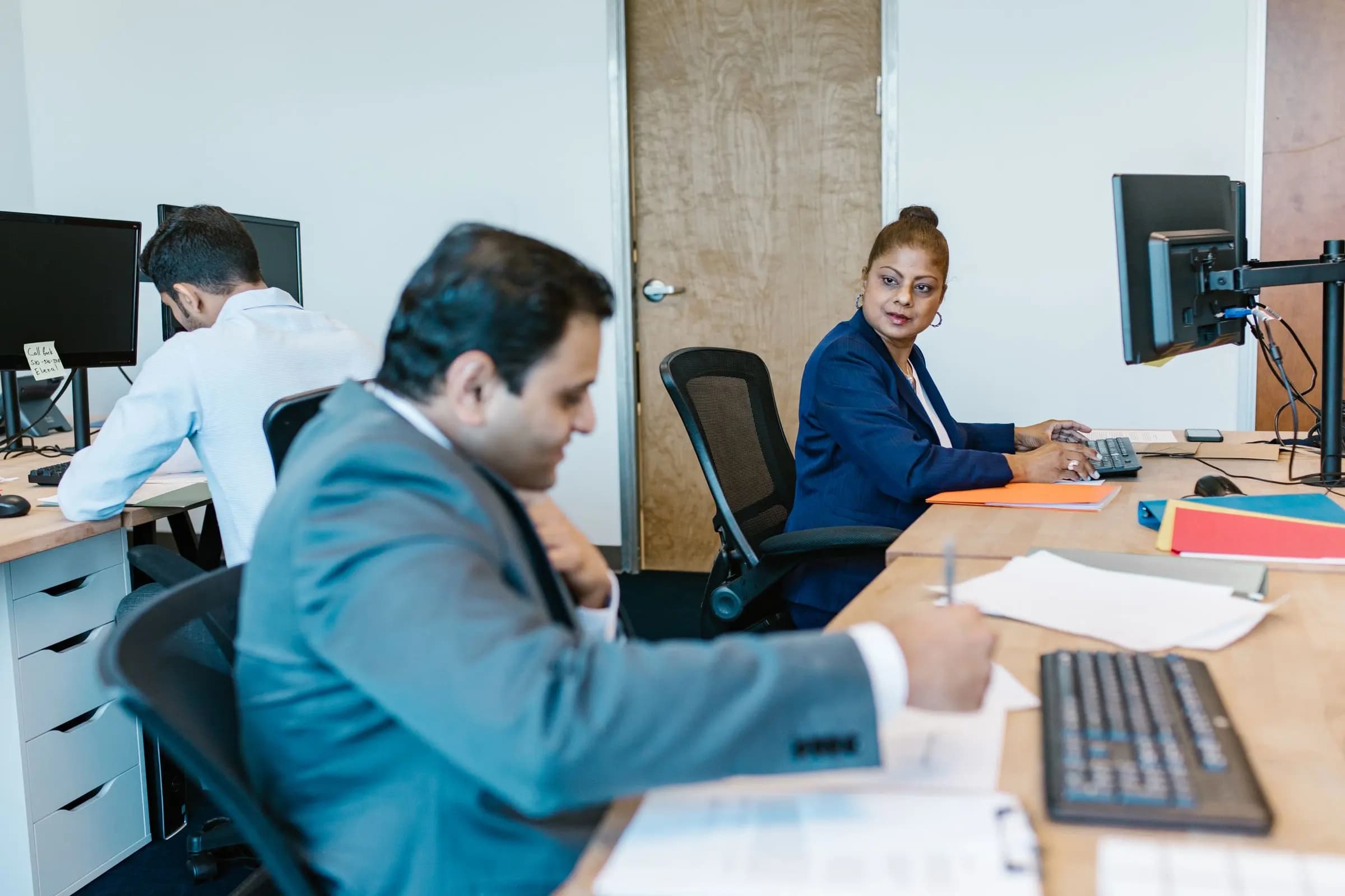 Colleagues work at their desks in an open office, one writing on paperwork while another looks over from her computer.