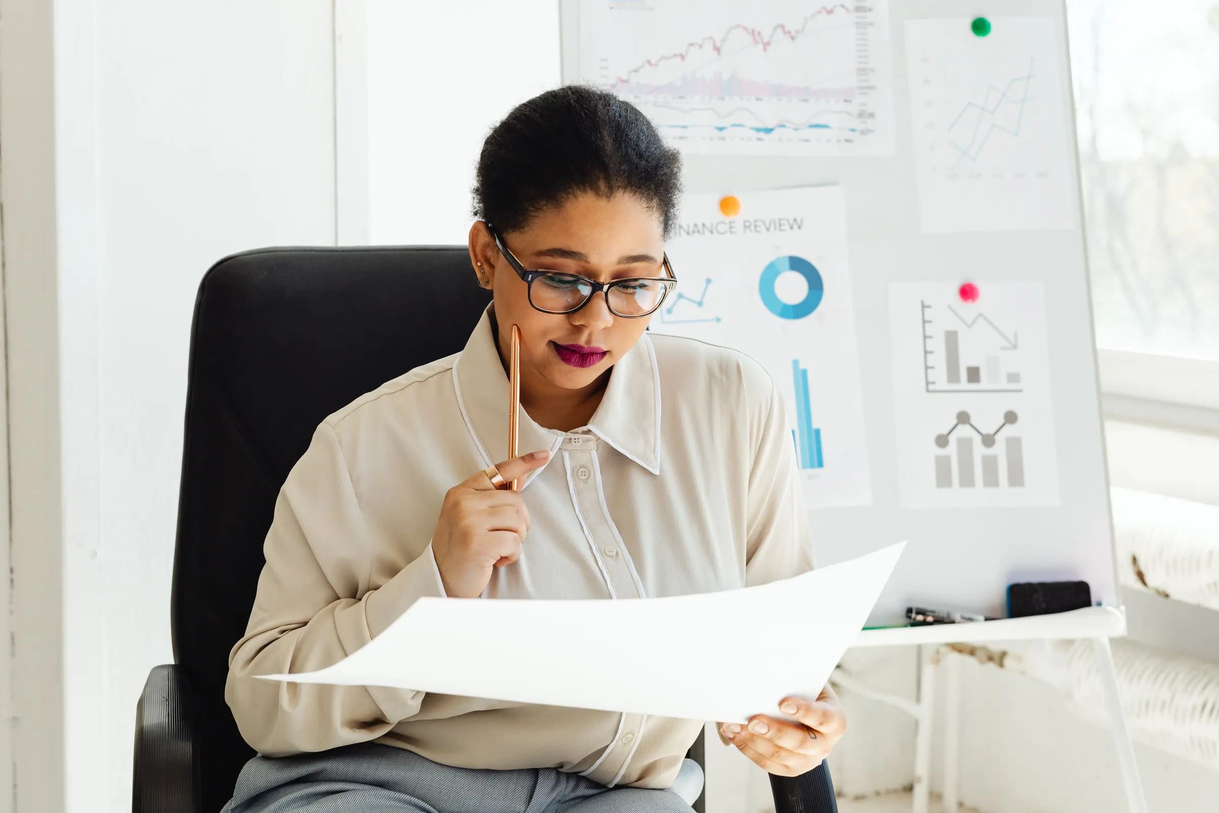 A woman in glasses studies a large printed sheet in front of a board pinned with charts.