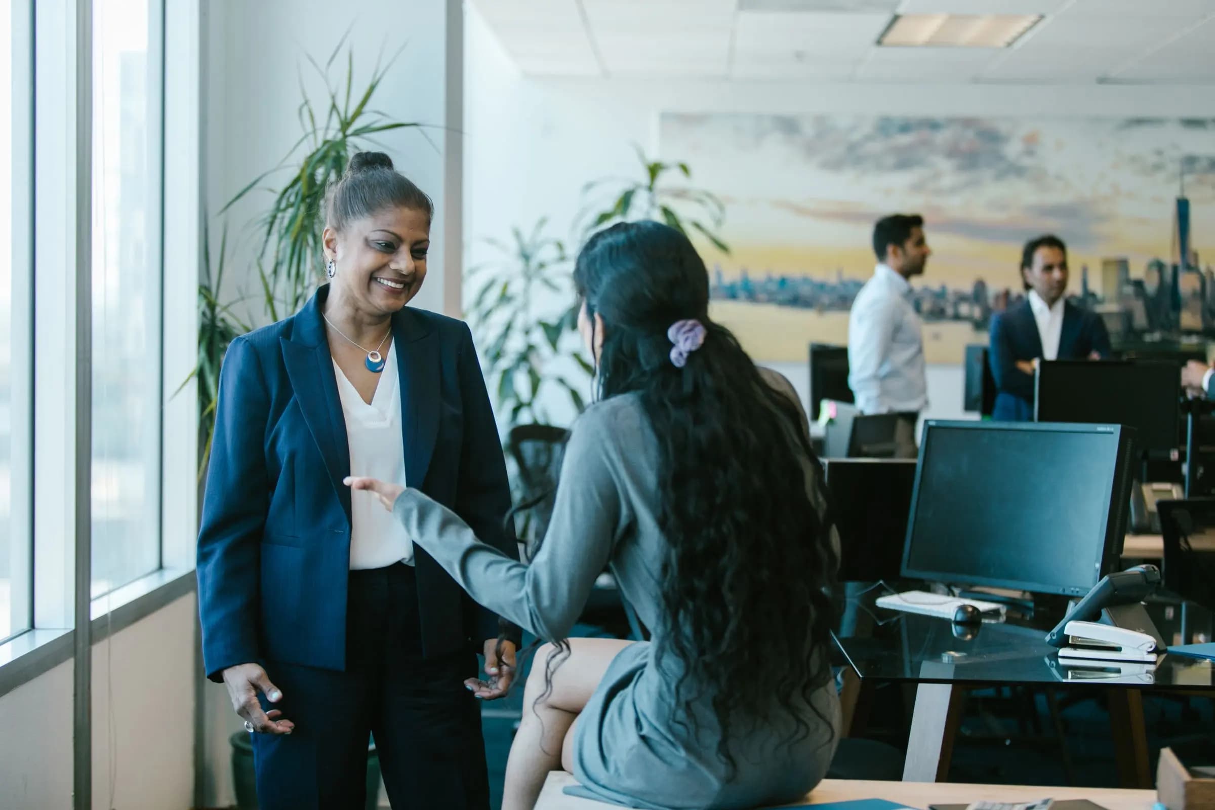 Two women chat by a window in an office while colleagues talk at desks in the background.
